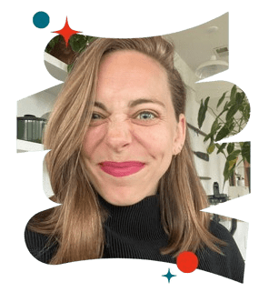 A smiling woman with medium-length hair and bright pink lipstick makes a playful face, wearing a black sweater, in a modern indoor setting with plants and shelves in the background.